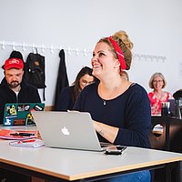 Studentin sitzt lachend vor dem Laptop im Seminarraum. Im Hintergrund eine sitzende Gruppe Studierender.