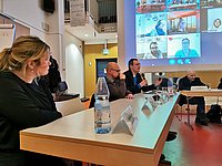Podiumsgäste der Veranstaltung sitzen nebeneinander an Tischen und reden. Im Hintergrund eine Leinwand auf der Personen zu sehen sind, die online teilnehmen. 