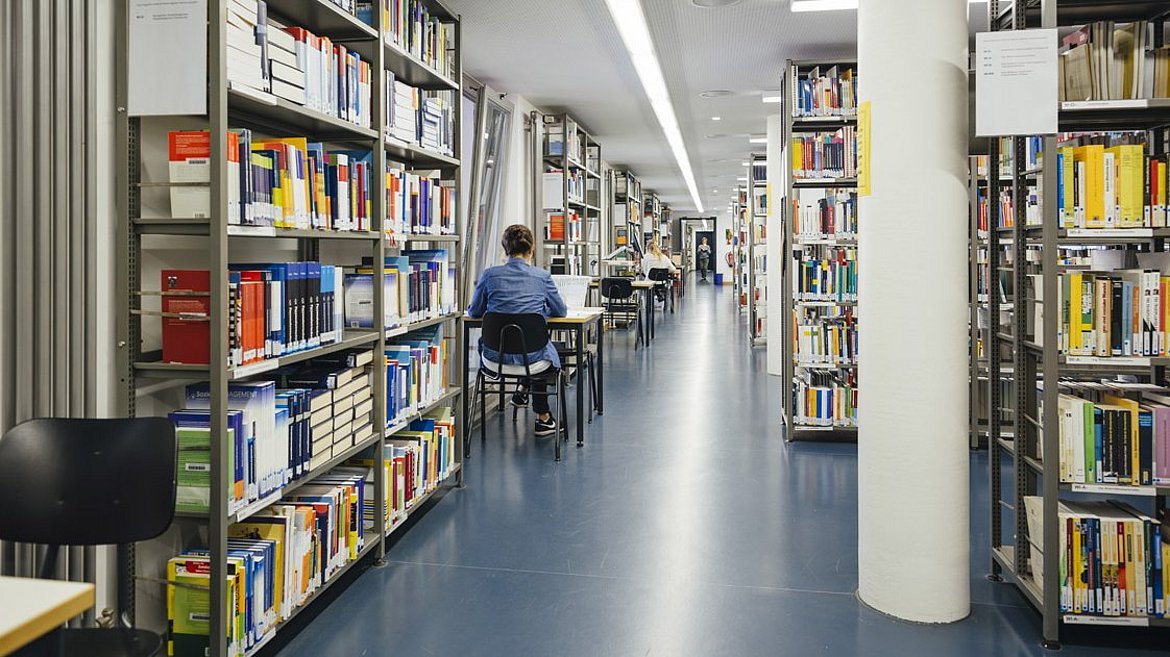 Blick durch die Bibliothek: geradeaus ein Gang, rechts Bücherregale, links sitzt eine Studentin an einem Tisch und arbeitet.