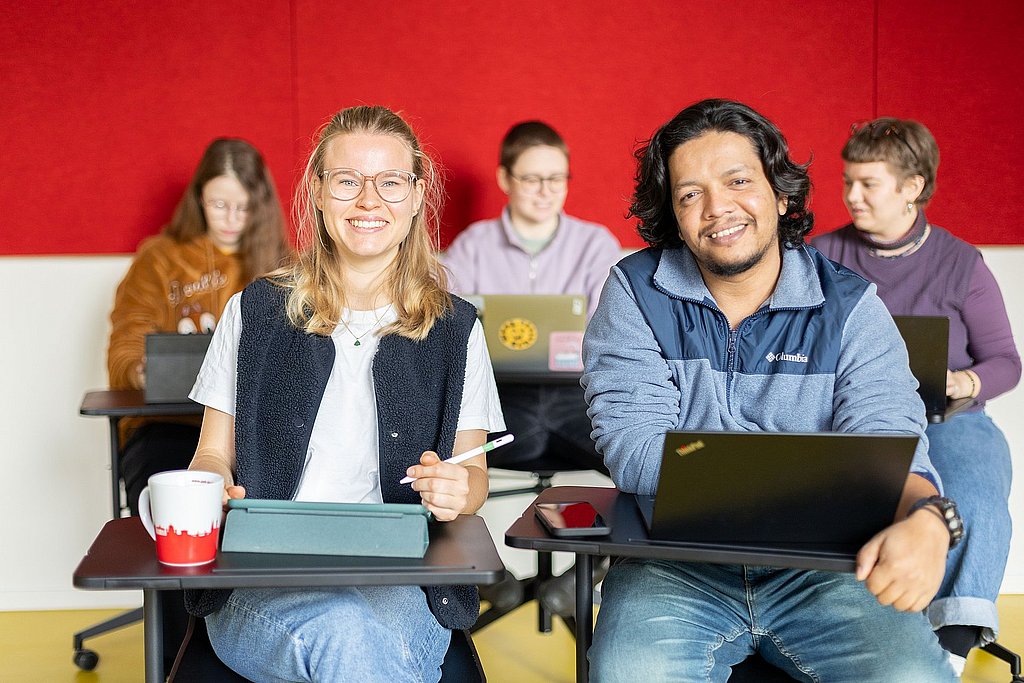 Fünf Studierende sitzen in einem Seminar der ASH Berlin. Zwei Studierende schauen lächelnd in die Kamera. 