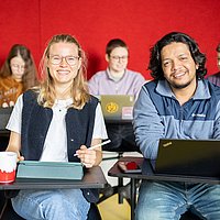 Fünf Studierende sitzen in einem Seminar der ASH Berlin. Zwei Studierende schauen lächelnd in die Kamera. 