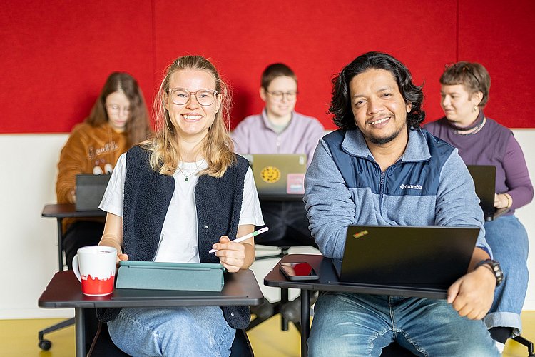 Fünf Studierende sitzen in einem Seminar der ASH Berlin. Zwei Studierende schauen lächelnd in die Kamera. 