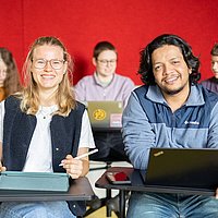 Fünf Studierende sitzen in einem Seminar der ASH Berlin. Zwei Studierende schauen lächelnd in die Kamera. 