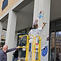 Das Foto zeigt die "MINIMAS", Hellersdorfer Maskotchen, die von Nachbar_innen gestaltet wurden, und hier an die Säulen vor der ASH Berlin geklebt werden.