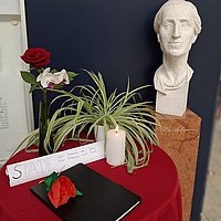 Auf dem Foto ist die Büste von Alice Salomon zu sehen, die sich im Foyer der ASH Berlin befindet, sowie ein Tisch mit Blumen, einer Trauerkerze und einem Kondolenzbuch
