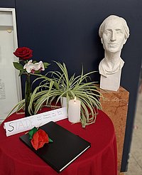 Auf dem Foto ist die Büste von Alice Salomon zu sehen, die sich im Foyer der ASH Berlin befindet, sowie ein Tisch mit Blumen, einer Trauerkerze und einem Kondolenzbuch