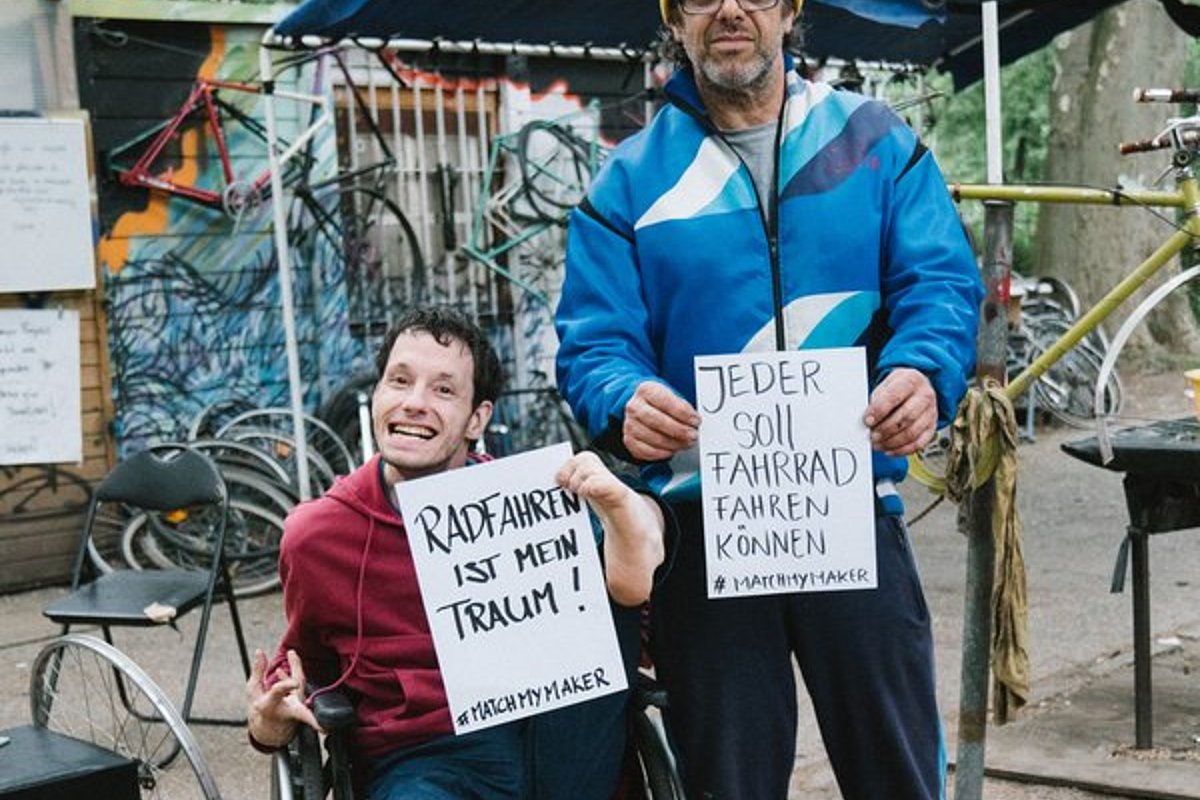 Eine Person im Rollstuhl hält ein Schild mit "Fahrrad fahren ist mein Traum". Daneben steht eine Person mit einem Schild "Jeder soll Fahrrad fahren können"