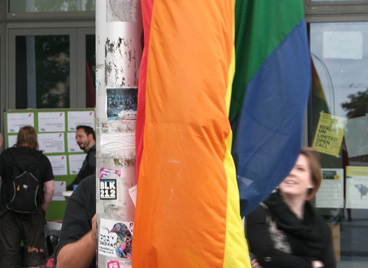 Detailansicht der Regenbogenflagge vor der ASH Berlin, dahinter verschwommen ein lachendes Gesicht