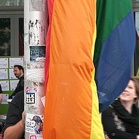 Detailansicht der Regenbogenflagge vor der ASH Berlin, dahinter verschwommen ein lachendes Gesicht