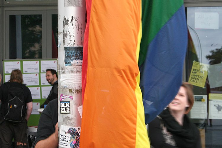 Detailansicht der Regenbogenflagge vor der ASH Berlin, dahinter verschwommen ein lachendes Gesicht