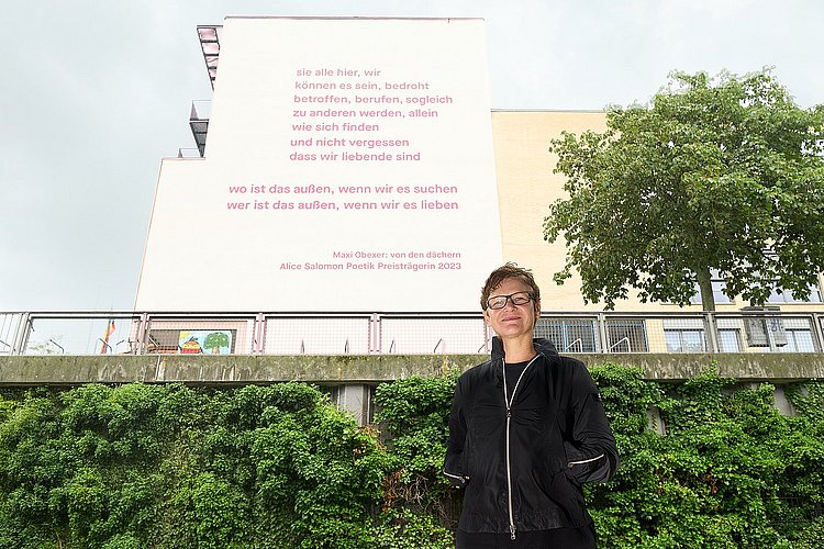 The photo shows Maxi Obexer in front of the south façade of ASH Berlin, on which Obexer's poem "von den dächern" can be read.
