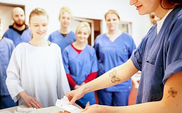 Eine Studentin simuliert einer Gruppe von Mitstudierenden im Skills Lab das Wickeln an einer Pflegepuppe
