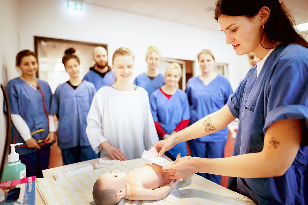 Eine Studentin simuliert einer Gruppe von Mitstudierenden im Skills Lab das Wickeln an einer Pflegepuppe