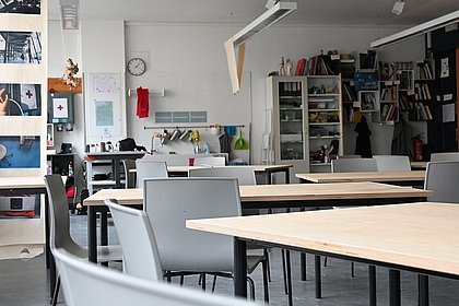 Es ist die Werkstatt mit Tischen und Stühlen mit Blick auf die Tür zu sehen. Im Hintergrund befinden sich Waschbecken und Bücherregal.