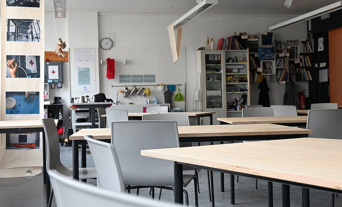 Auf dem Bild sind Tische und Stühle, im Hintergrund das Waschbecken und die Büchersammlung zu sehen.