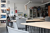 Es ist die Werkstatt mit Tischen und Stühlen mit Blick auf die Tür zu sehen. Im Hintergrund befinden sich Waschbecken und Bücherregal.