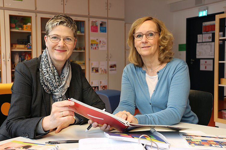 Corinna Schmude und Anja Voss sitzen an einem Besprechungstisch und halten das Berliner Bildungsprogramm für Kitas und Kindertagespflege in den Händen