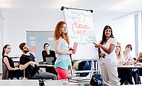 Studierende stehen in einem Seminarraum vor einem Flipchart und halten vor ihren Kommiliton_innen einen Vortrag.