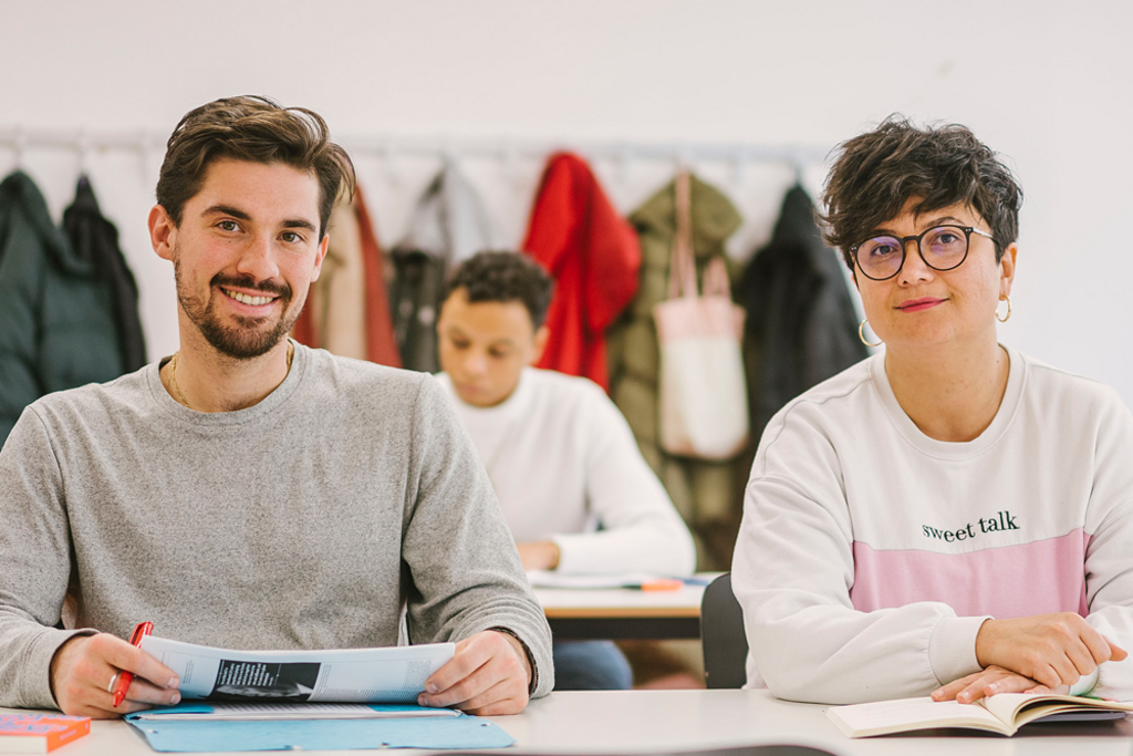 Auf dem Foto sind zwei Studierende der ASH Berlin zu sehen. Sie sitzen an einem Tisch in einem Seminarraum und schauen den Betrachter_innen in die Augen.