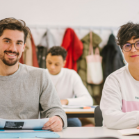 Auf dem Foto sind zwei Studierende der ASH Berlin zu sehen. Sie sitzen an einem Tisch in einem Seminarraum und schauen den Betrachter_innen in die Augen.