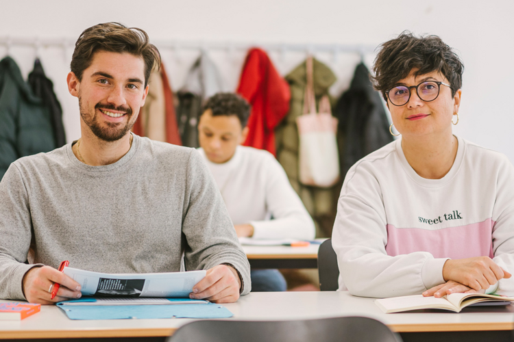 Auf dem Foto sind zwei Studierende der ASH Berlin zu sehen. Sie sitzen an einem Tisch in einem Seminarraum und schauen den Betrachter_innen in die Augen.