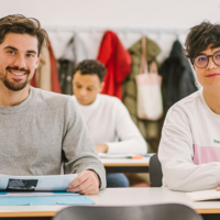 Auf dem Foto sind zwei Studierende der ASH Berlin zu sehen. Sie sitzen an einem Tisch in einem Seminarraum und schauen den Betrachter_innen in die Augen.