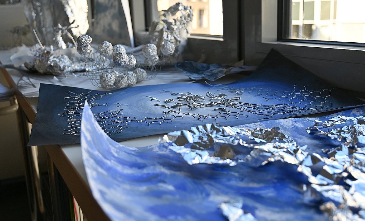 Zu sehen sind nebeneinander liegende Werke auf dessen blauen Papieruntergrund verschiedene silbern-metallische Materialien liegen.