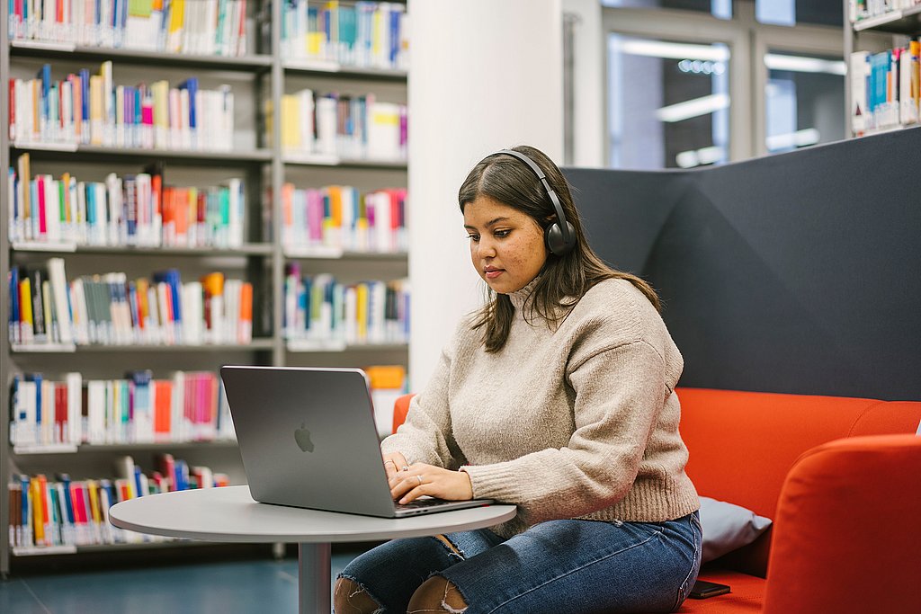 Eine Studierende mit Headset sitzt an einem Laptop in einer Bibliothek.