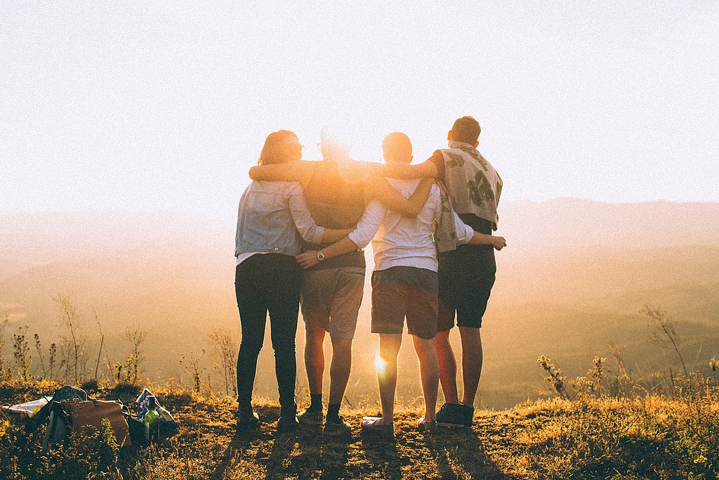 Four people with their arms around each other's shoulders stand on a meadow and gaze into the sunset. They have their backs to the camera.