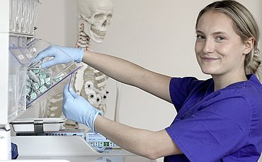 Eine Studierende entnimmt im Skillslab Instrumente für die Pflege. Sie trägt medizinische Handschuhe und lächelt in die Kamera.