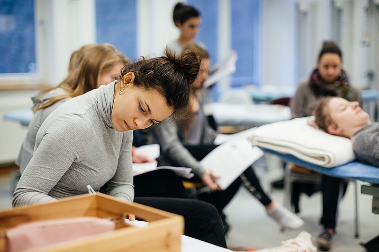 Eine Studentin des primärqualifizierenden Studiengangs macht sich Notizen. Im Hintergrund sind weitere Studierende zu sehen, die um eine Liege herum sitzen.