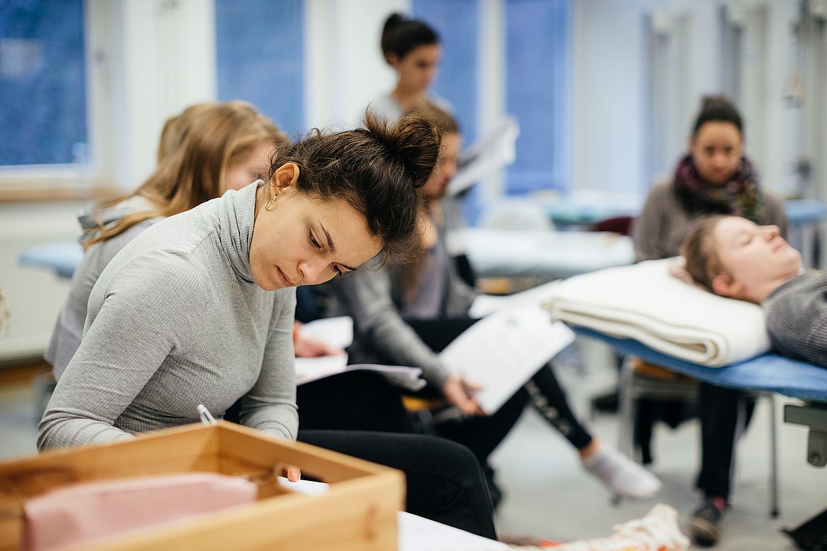 Eine Studentin des primärqualifizierenden Studiengangs macht sich Notizen. Im Hintergrund sind weitere Studierende zu sehen, die um eine Liege herum sitzen.