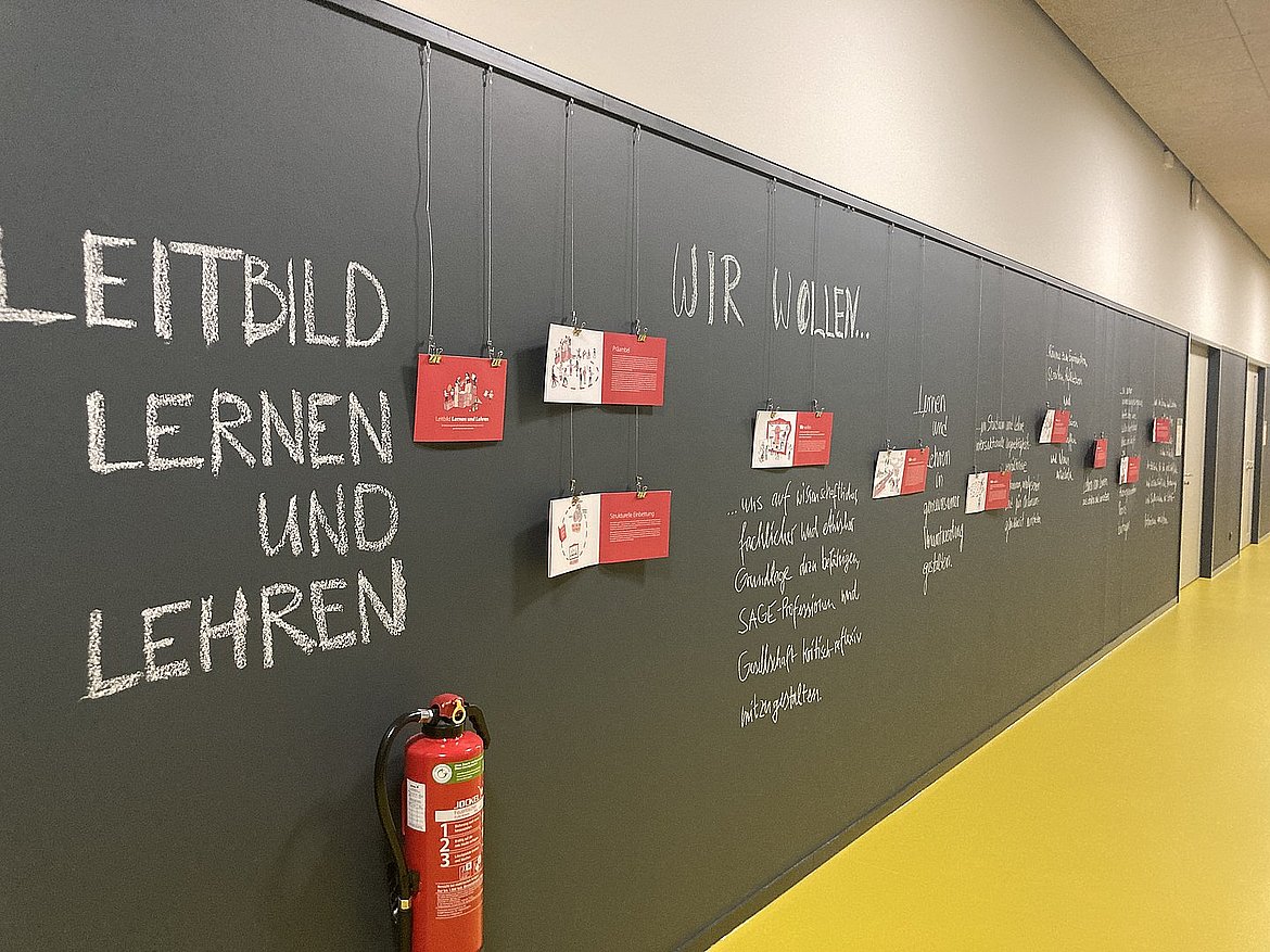 Flur mit einer Wandtafel. Darauf steht in weißer Kreide ‚Leitbild Lernen und Lehren‘, ergänzt durch weitere Notizen und aufgehängte rote Karten mit Informationen
