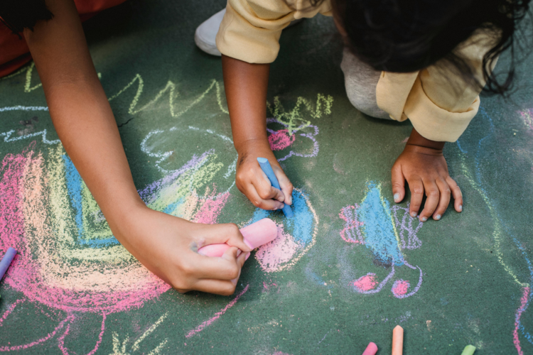 Von oben fotografiert: ein Kind, das auf der Straße mit Kreide malt, daneben eine erwachsenere Hand