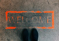 The photo shows the fading lettering Welcome sprayed in orange on the street. Two feet can be seen at the bottom, as if the person is looking at the lettering.