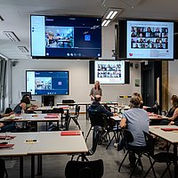 Teilnehmende des Alumnitags sitzen in einem Seminarraum an der ASH Berlin. Sie sitzen an Seminartischen und hören dem Vortrag einer Rednerin zu. 