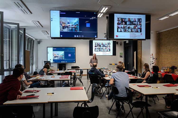 Teilnehmende des Alumnitags sitzen in einem Seminarraum an der ASH Berlin. Sie sitzen an Seminartischen und hören dem Vortrag einer Rednerin zu. 
