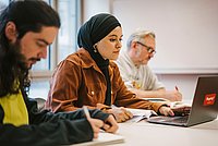 Drei Studierende sitzen nebeneinander in einem Seminarraum und arbeiten am Laptop oder schreiben sich Notizen auf.