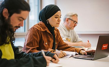 Drei Studierende sitzen nebeneinander in einem Seminarraum und arbeiten am Laptop oder schreiben sich Notizen auf.