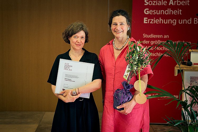 Yevgenia Belorusets und Bettina Völter auf der Bühne des Audimax der ASH Berlin. Belorusets hält die Urkunde in den Händen und Völter trägt einen kleinen Olivenbaum.