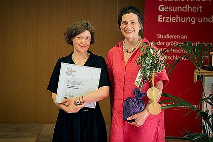Yevgenia Belorusets und Bettina Völter auf der Bühne des Audimax der ASH Berlin. Belorusets hält die Urkunde in den Händen und Völter trägt einen kleinen Olivenbaum.