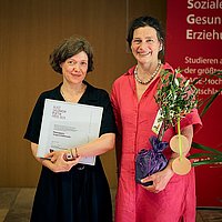 Yevgenia Belorusets und Bettina Völter auf der Bühne des Audimax der ASH Berlin. Belorusets hält die Urkunde in den Händen und Völter trägt einen kleinen Olivenbaum.