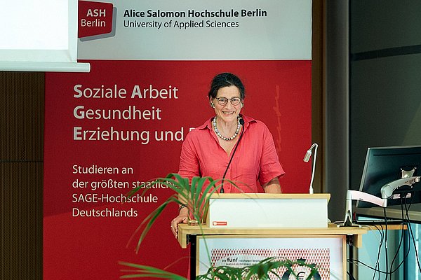 Bettina Völter auf der Bühne im Audimax der ASH Berlin. Sie steht hinter dem Rednerpult und begrüßt die Gäste.