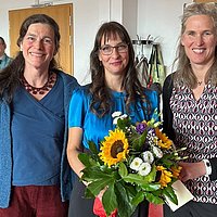 Gruppenfoto von Bettina Völter, Jana Einsporn und Gesine Bär. Die drei stehen nebeneinander im Audimax der ASH Berlin. Jana Einsporn hält einen Blumenstrauß in der Hand.