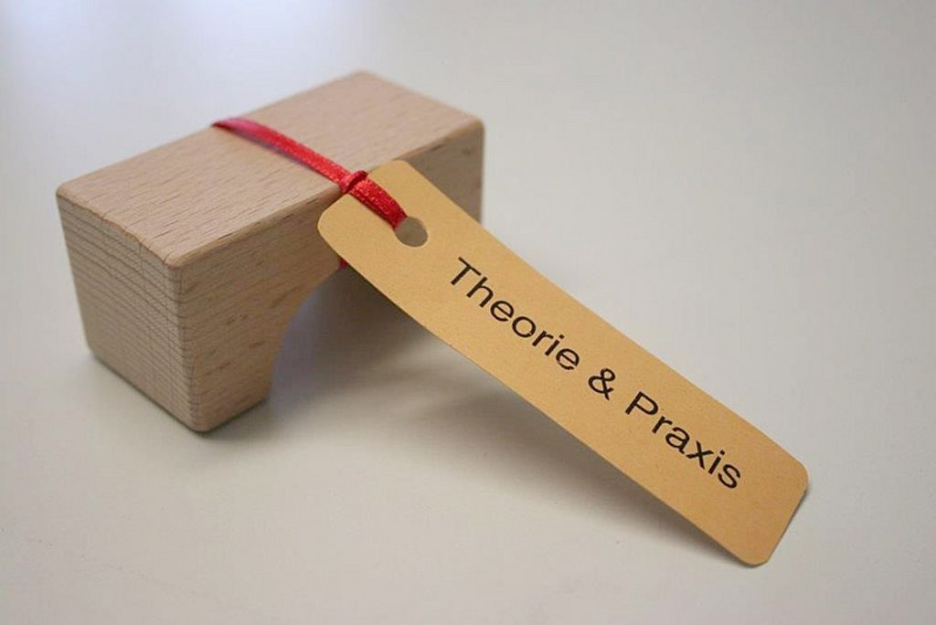 An einem Bauklotz aus Holz ist ein kleines Schild mit der Aufschrift Theorie und Praxis befestigt.