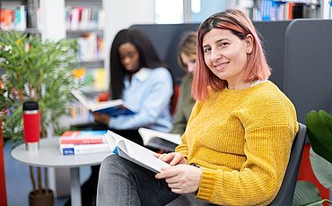 Ein Studierende mit einem Buch auf dem Schoß schaut, in der Bibliothek sitzend, lächelnd in die Kamera. Im Hintergrund sind zwei Studierende zu sehen.