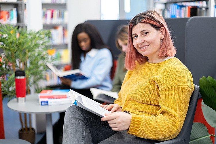 Ein Studierende mit einem Buch auf dem Schoß schaut, in der Bibliothek sitzend, lächelnd in die Kamera. Im Hintergrund sind zwei Studierende zu sehen.