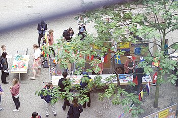 Das Bild zeigt eine bunte Großaufnahme des International Day. Man sieht Tische, Bäume und Gäste des International Day.