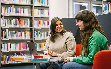 Zwei Studierende sitzen in der Bibliothek und unterhalten sich. Vor ihnen ein Tisch mit Büchern. Im Hintergrund stehen Bücherregale.