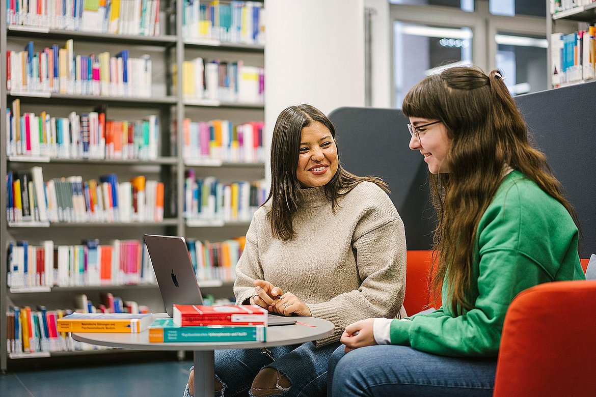 Zwei Studierende sitzen in der Bibliothek und unterhalten sich. Vor ihnen ein Tisch mit Büchern. Im Hintergrund stehen Bücherregale.
